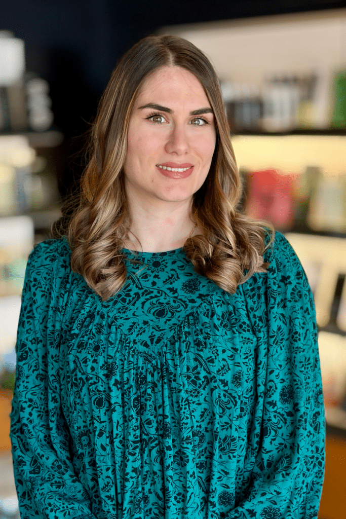 Woodhaven hair stylist specializing in highlights, dimensional blonding and precisions haircuts standing in front of an Aveda haircare display of home care products